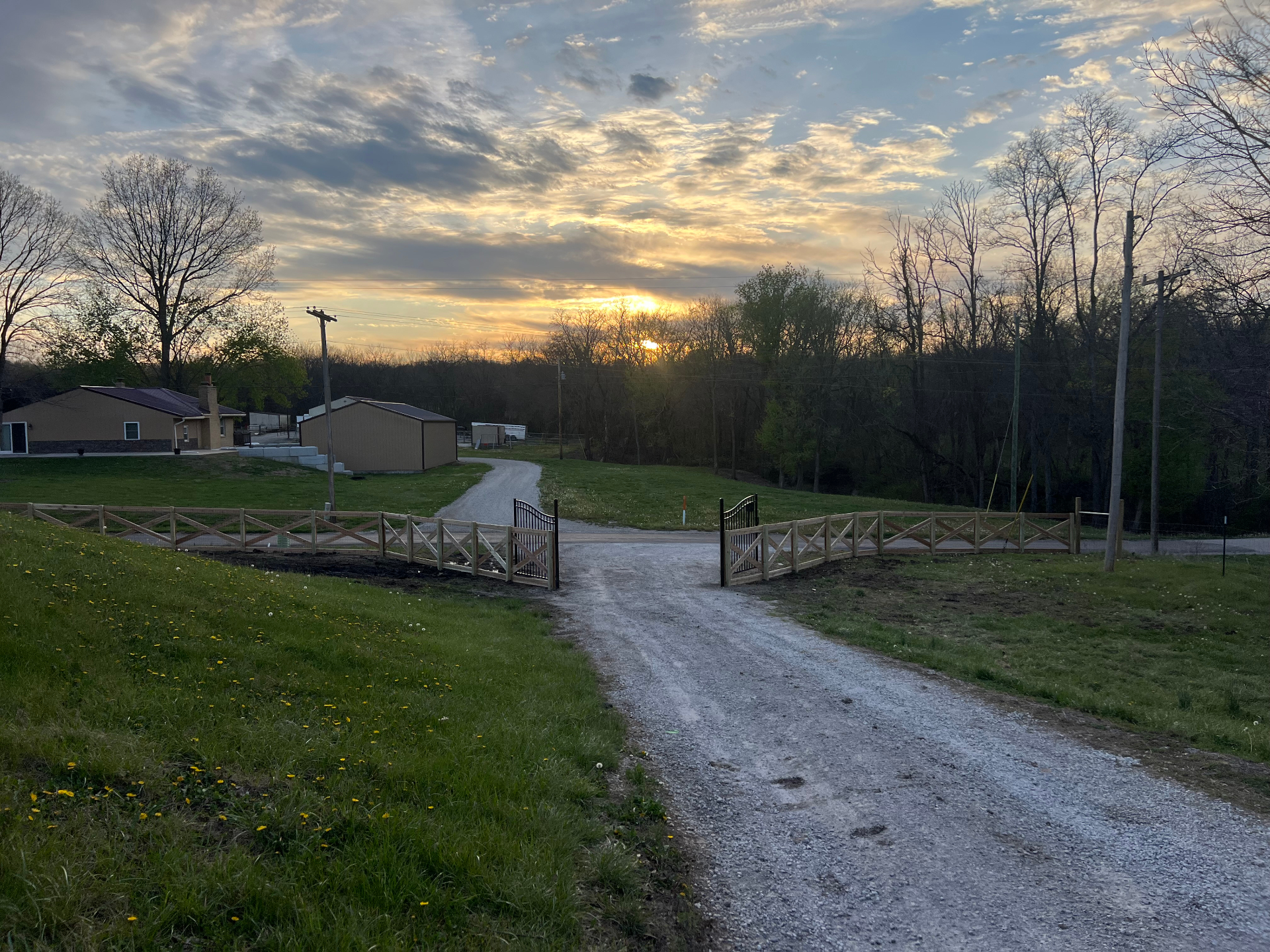 Sunset driveway view of red cedar cross rail ranch fence with custom black steel double arch entry gates and walk gate — Spring Hill, KS